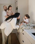 A family enjoys time together in a bright kitchen. The mother smiles at her baby, who sits on the counter atop a mushie Extra Soft Muslin Changing Pad Cover, while the father holds their daughter. Everyone looks happy and engaged.