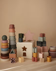 A set of mushie Stacking Cups Toy and other colorful Danish Hygge toys, including rings, blocks, and a shape sorter with star and cross-shaped holes, are displayed on a neutral beige background.