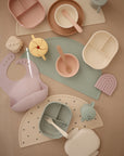 Pastel mushie silicone baby feeding items, including divided plates, bowls, cups, a bib, utensils, placemats, and the non-toxic mushie Lunchbox in playful rainbow and bear shapes, are neatly displayed on a beige background.