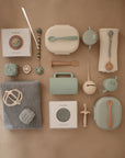 A flat lay of pastel baby items on a beige background, featuring the mushie Lunchbox with compartments, non-toxic plastic pacifiers, containers, teether, rattle, spoon, pacifier clip, bib, and muslin cloth.