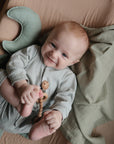 A smiling baby with light hair lies on a mushie Extra Soft Muslin Changing Pad Cover, wearing a light outfit, holding a beaded wooden toy, grabbing their foot, and partially covered by a green blanket.