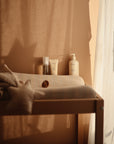A softly lit baby changing table with a mushie Extra Soft Muslin Changing Pad Cover, star-shaped pillow, pacifier, and baby skincare bottles. Sheer curtains cast gentle shadows on the peach-colored wall.