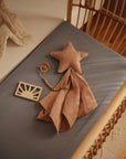 A cozy crib with a blue mattress sheet holds the mushie Star Lovey Blanket, a pink cloth, a beaded pacifier clip, and a wooden sun toy. Warm lighting creates a soft, calming atmosphere.