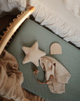 A cozy nursery featuring a woven crib, mint green mattress, star-shaped pillow, wooden rainbow toy, and the mushie Star Lovey Blanket made from organic cotton, all set beside a white ruffled bedspread.