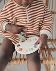A young child in a red-and-white striped outfit sits on a rattan chair, holding a pastel sensory toy and has a FRIGG Daisy Natural Rubber Pacifier from the FRIGG Daisy Natural Rubber Pacifier 2-Pack clipped to their shirt.
