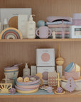 Two wooden shelves display pastel-colored baby products, including mushie bottles, cups, plates, stacking toys, silicone bibs, the Flower Training Toothbrush by mushie, and skincare items—all organized in soft muted tones.
