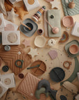 A flat lay of pastel mushie baby feeding items—divided plates, bowls, cups, bibs, teething rings, soft toys—and the mushie Star Lovey Blanket in organic cotton, all arranged with packaging on a cream surface with quilted mats and star shapes.