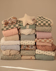 Three neat stacks of patterned and solid-colored fabrics in soft, muted tones. Atop the center stack is a star-shaped pillow. The mushie Extra Soft Muslin Changing Pad Cover adds comfort to the calm, beige backdrop.
