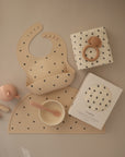 A flat lay of beige baby feeding items with black floral patterns from mushie, including a silicone bib, bowl with spoon, sippy cup, teether, pacifier, muslin washcloth, and Organic Cotton Muslin Swaddle Blanket.