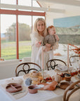 A woman holding a baby stands by a dining table set for a meal with mushie Round Dinnerware Plates, autumn-themed decor, and glasses in a bright room with large windows overlooking nature.