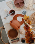 A toddler meal table set with earth-tone silicone plates, bowls, utensils, bibs, and the FRIGG Andersen Fairytale Natural Rubber Pacifier 2-Pack by FRIGG, surrounded by autumn leaves, a candle, and festive décor on a white tablecloth.