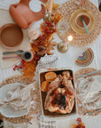 A Thanksgiving table set with woven placemats, mushie Round Dinnerware Plates, napkins, candles, and autumn leaves features a roasted chicken with orange slices in a baking dish at the center.