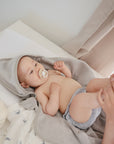 A baby with a pacifier, wrapped in a gray hooded towel, lies on a mushie Extra Soft Muslin Changing Pad Cover while an adult kisses their feet. Bottles and baby products sit nearby.