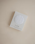 A mushie box displays the Extra Soft Muslin Changing Pad Cover, with its white textured fabric visible through a circular window on the packaging, all set against a neutral beige background.