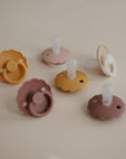 Six FRIGG Daisy Silicone Pacifier 2-Packs by FRIGG—made from medical grade silicone—feature scalloped edges, round nipples, and circular handles in muted brown, beige, pink, and mustard shades for ultimate baby comfort.
