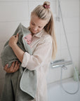 A woman in a bathroom, wearing a light striped shirt and pink scrunchie, holds a baby wrapped in a green towel as she prepares to apply mushie Green Lemon Baby Body Lotion. White tiles and toiletries are visible in the background.