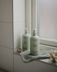 Two pale green bottles of mushie Green Lemon Baby Body Lotion sit on a soft towel beside a window, with a pacifier and coral nearby, all arranged on the bathroom's tiled windowsill.