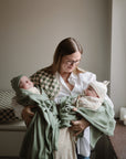 A woman with glasses smiles while holding two babies wrapped in green blankets and mushie Organic Cotton Muslin Burp Cloths (2-Pack) in a softly lit room near a window.