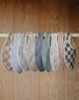 Nine mushie Muslin Bibs in assorted colors and patterns—checks, paisley, solids, and florals—are neatly displayed on a wooden rod with a wood background.