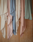The mushie Stretchy Swaddle scarves hang against a wooden wall, draping in soft folds. Available in cream, blush pink, blue, and green—some with subtle patterns and others solid—they're crafted for breathability like a baby blanket.