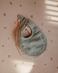 Two mushie Muslin Bibs, one in sage green and one in light pink, are stacked on a peach fabric with an orange rainbow pattern. Window-blind light creates soft shadows across the scene.
