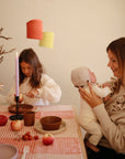 A woman smiles while holding a baby at a candlelit table with fruit and festive decor. Beside them, a young girl cuts food on mushie Round Dinnerware Plates. The scene has a warm, cozy atmosphere with soft lighting.