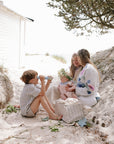 A woman and three kids picnic with a mushie Diaper Caddy on sand near a white cabin and trees.