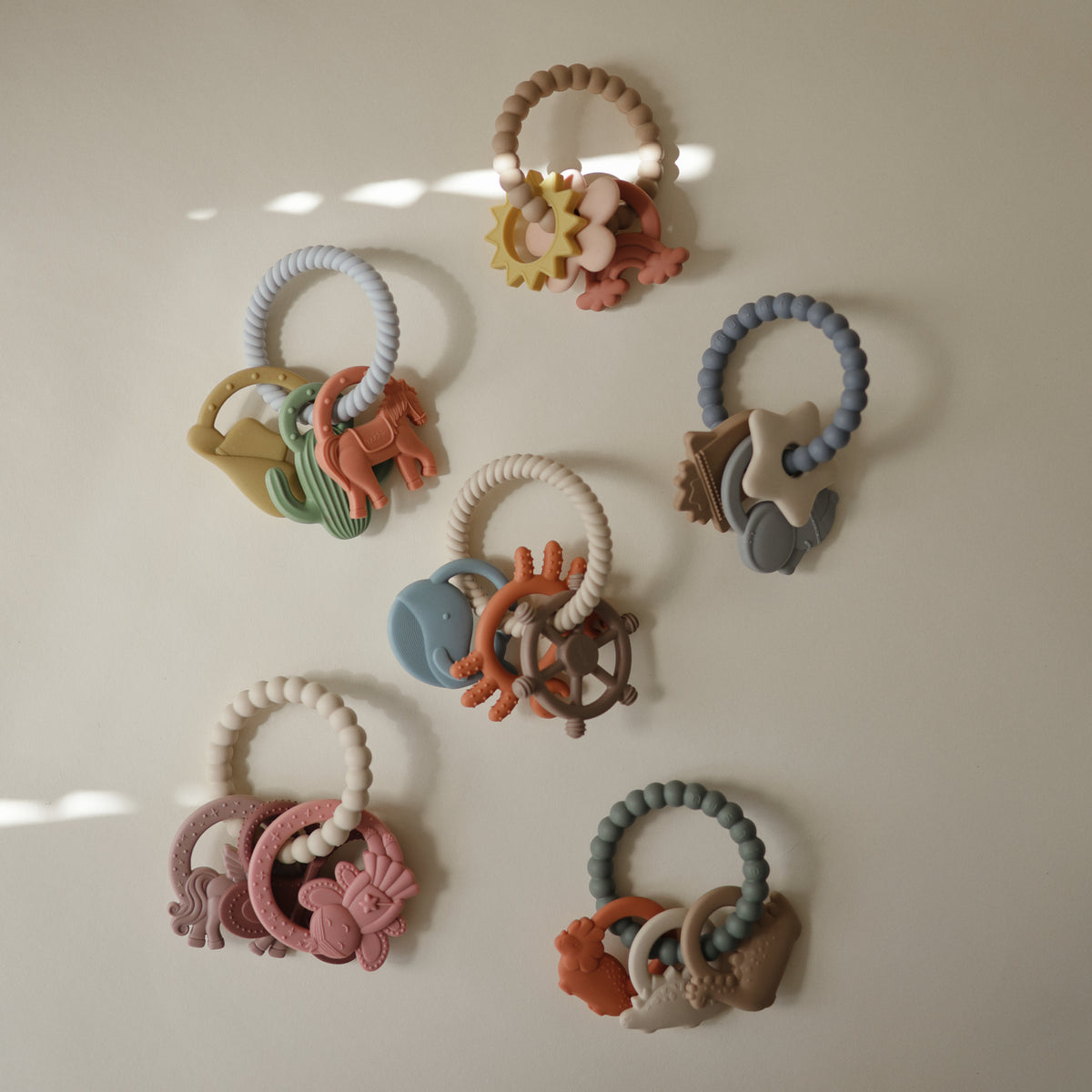 Five mushie Teething Rings with various charms arranged on a light surface.