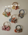 Five mushie Teething Rings with various charms arranged on a light surface.
