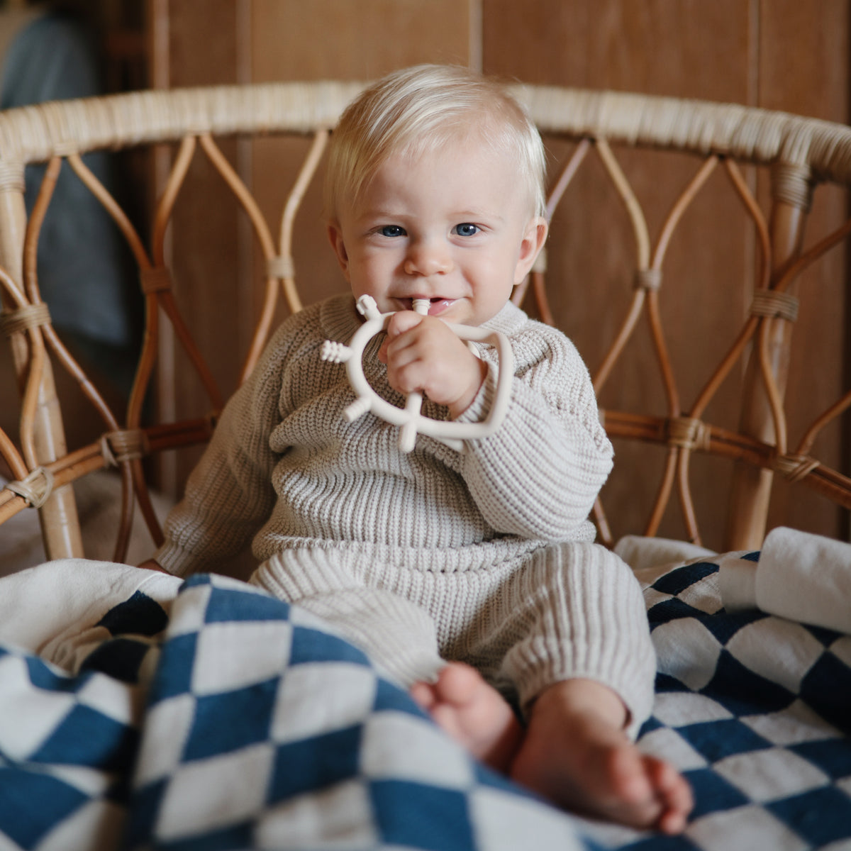 Baby in beige holds mushie Teething Gift Set, blue-checkered blanket by wicker chair.
