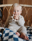Baby in beige holds mushie Teething Gift Set, blue-checkered blanket by wicker chair.
