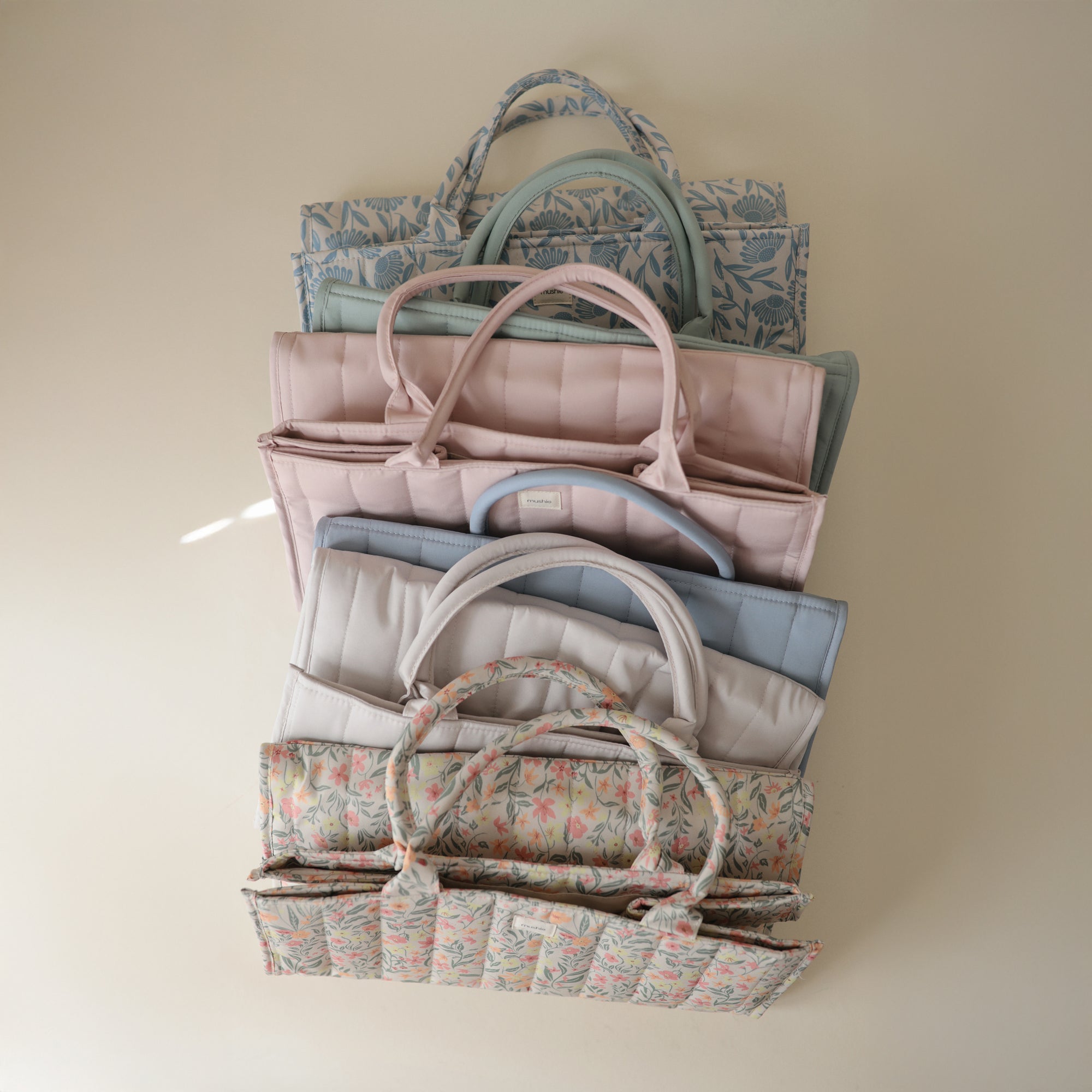 Lifestyle image of a stack of mushie Diaper Caddies in various colors and patterns on a beige background.