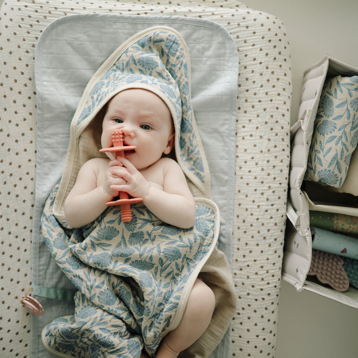Baby on changing pad with blue floral towel, teether, and mushie Diaper Caddy nearby.