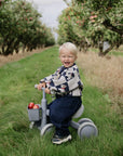A toddler riding a small gray mushie Mini Toddler Bike filled with apples in an orchard.