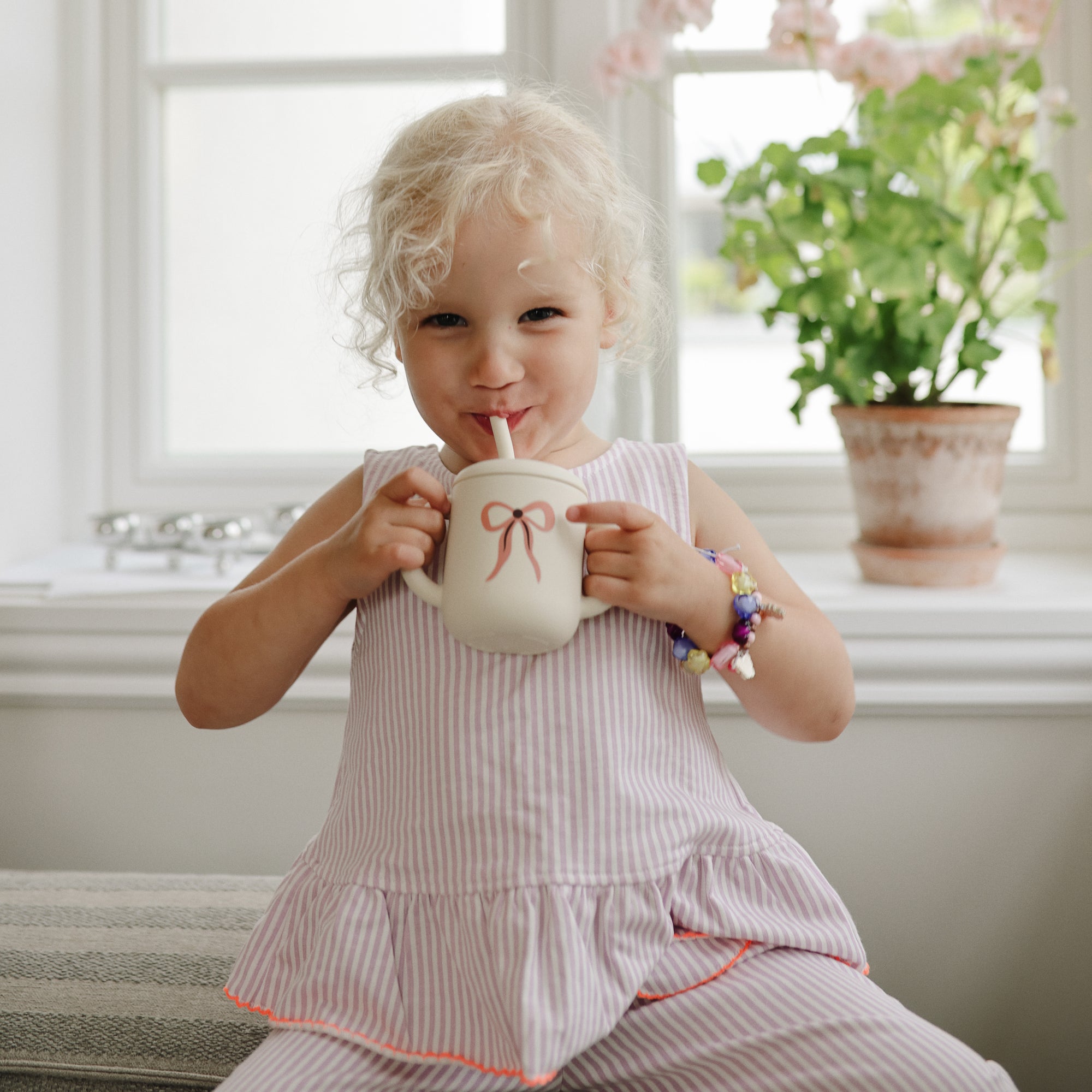 Happy child drinks from Mushie Silicone Training Cup + Straw by a window with plants.