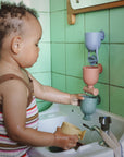 A toddler explores with mushie's Pour and Play Bath Set in a green-tiled bathroom sink.