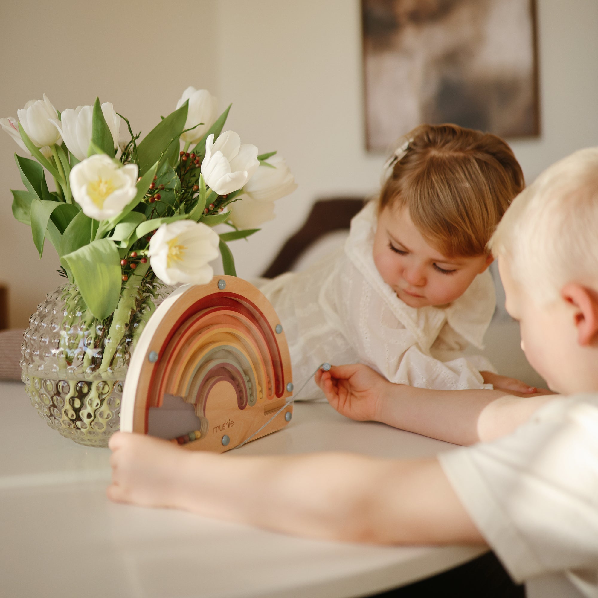Two children enjoy screen-free play with the mushie Wooden Magnetic Rainbow Maze on a table next to white tulips. The girl watches as the boy interacts with the toy, supporting creativity and fine motor skills in a cozy setting.