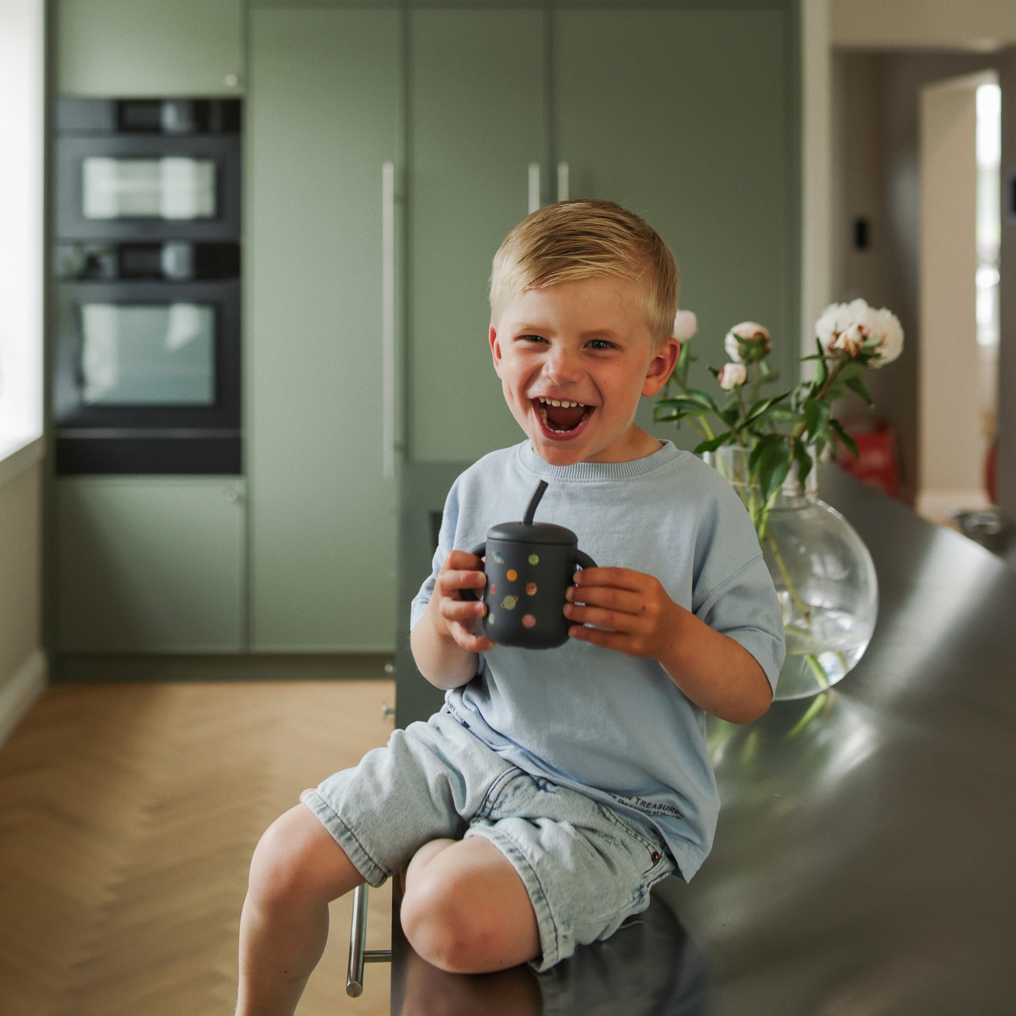 Smiling boy in blue shirt holds a Mushie Silicone Training Cup + Straw on the kitchen counter.