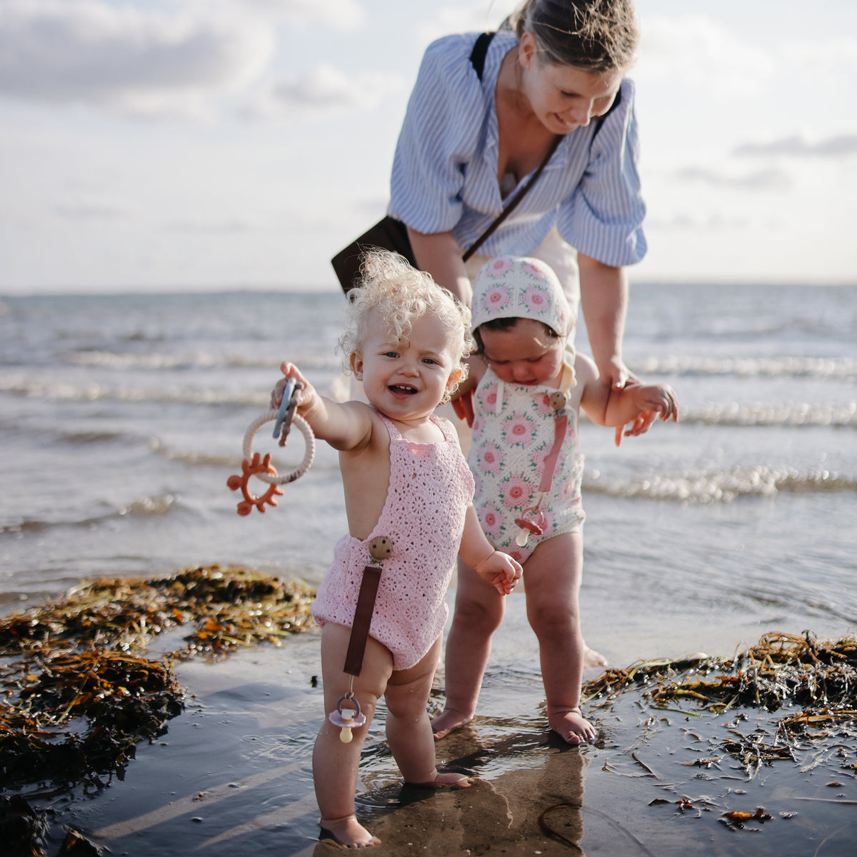 A toddler stands in shallow water holding a sun-shaped mushie Teething Ring.
