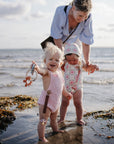 A toddler stands in shallow water holding a sun-shaped mushie Teething Ring.