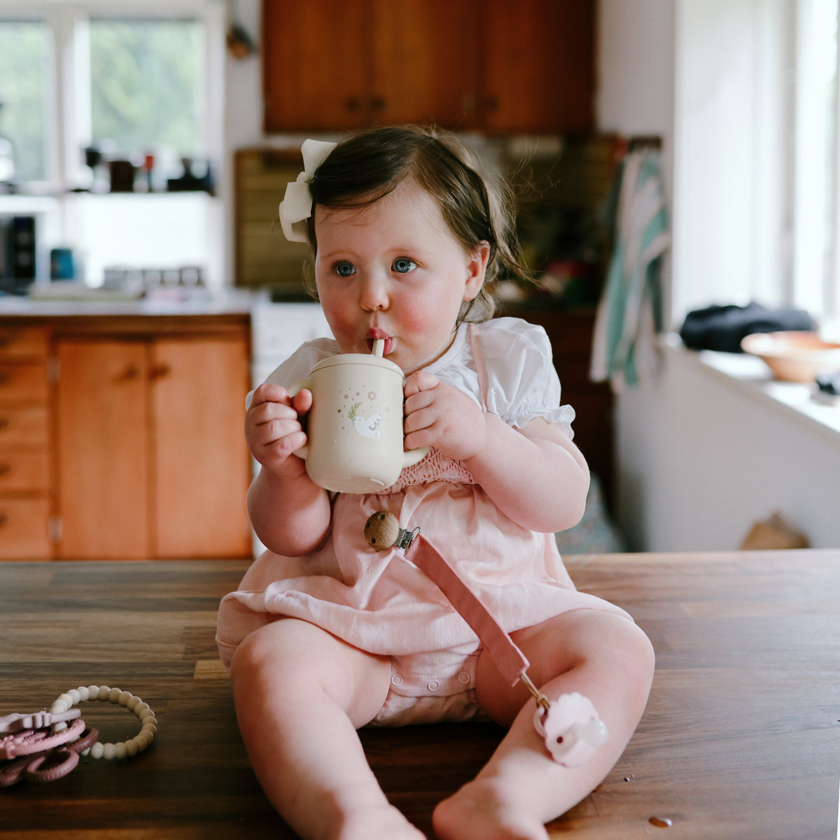 Toddler in pink uses Mushie Silicone Training Cup + Straw on the kitchen counter.