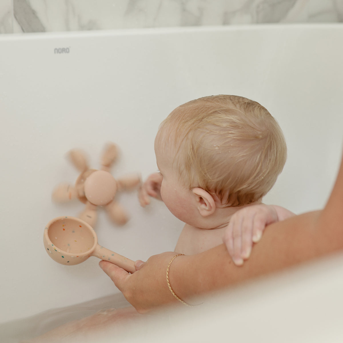 Baby playing with mushie Water Wheel Bath Toy in a tub, adult holds scooping cup nearby.