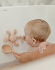 Baby playing with mushie Water Wheel Bath Toy in a tub, adult holds scooping cup nearby.