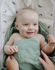 A smiling baby wrapped in sage green muslin on a Mushie Changing Pad Liner.