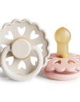 Product image of two Frigg Andersen Rubber baby pacifier, one Cream with heart designs and the other Blush, on a white background.
