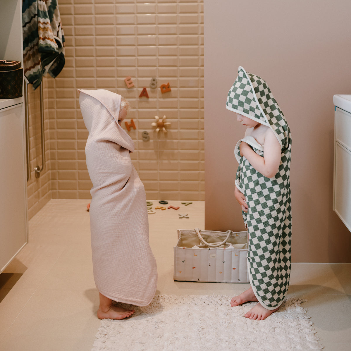 Two kids in hooded towels stand by a mushie Diaper Caddy in the bathroom.