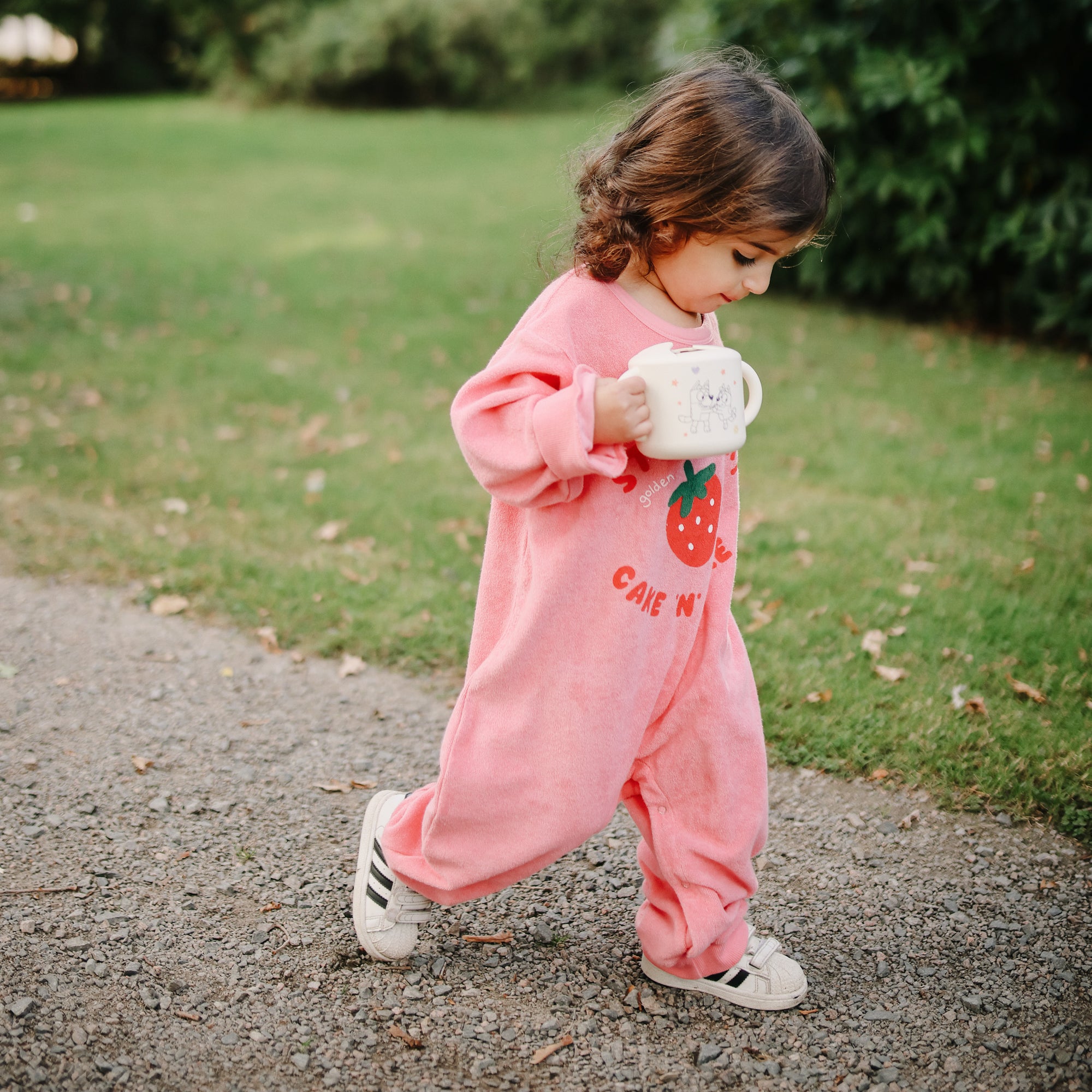 Toddler in pink onesie walks outside holding a mushie Snack Cup in both hands.