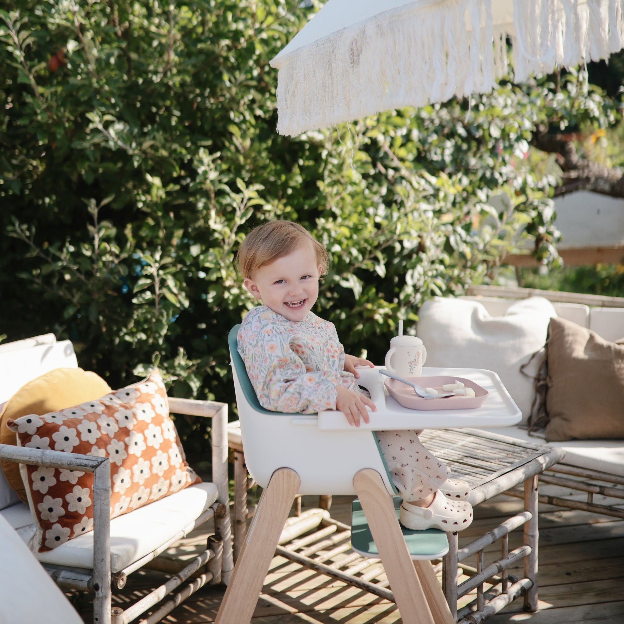 Smiling toddler outdoors, sipping from Mushie Silicone Training Cup + Straw in high chair.