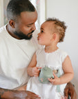 A man and toddler smile indoors while using the Mushie Silicone Training Cup + Straw.