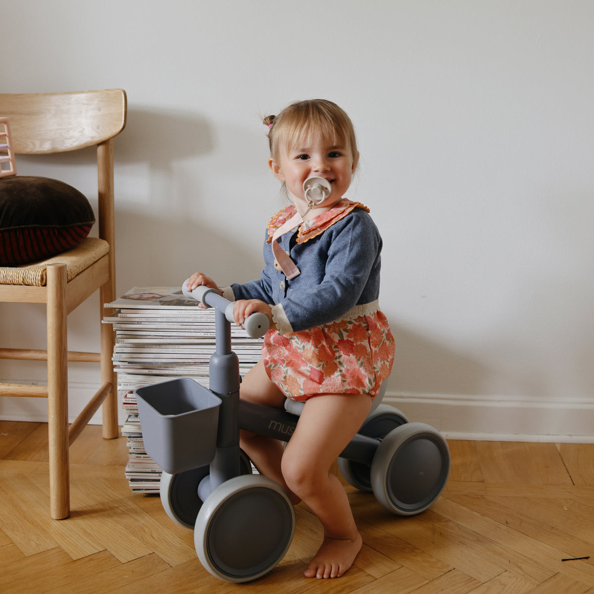 A toddler sits on a gray three-wheeled mushie Mini Toddler Bike with a front basket.